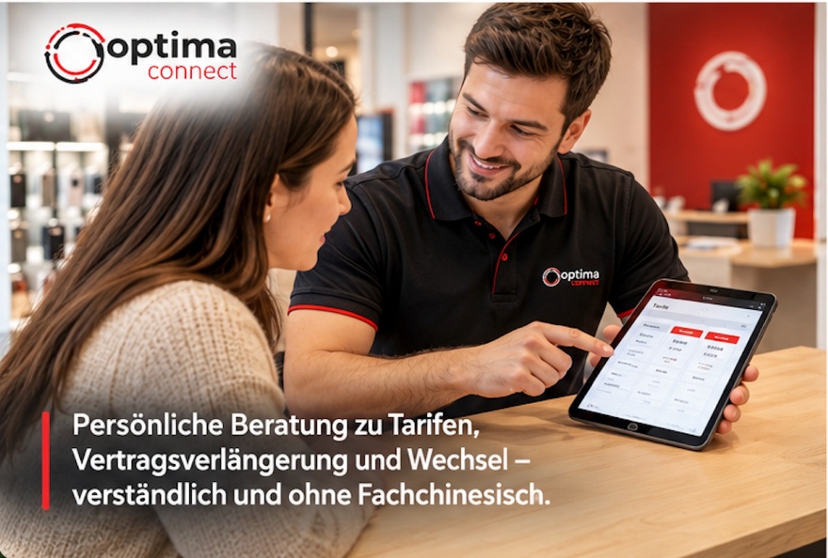 Ein lächelnder Mitarbeiter in schwarzem Poloshirt zeigt einer Kundin auf einem Tablet Informationen, während sie an einem Holztisch in einem modernen Büro mit rotem Akzent sitzen.